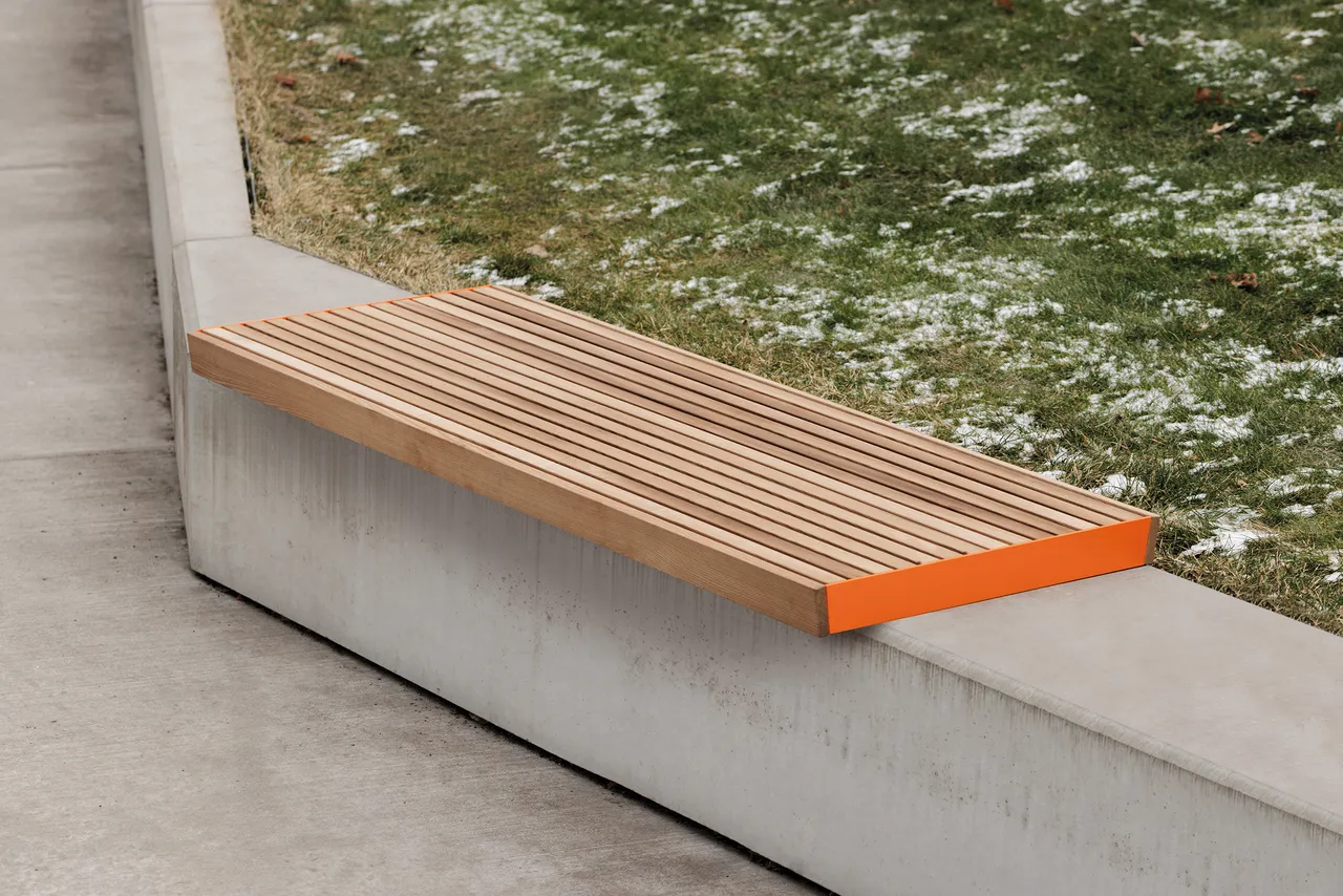 Die Abb. zeigt die EGOÉ Bank A1 zur Sockelmontage ohne Rückenlehne, mit Holz aus Thermoesche Natur und Gestell in Hellrotorange (RAL 2008).